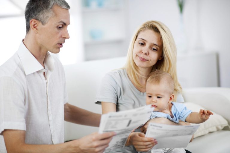 A worried couple looking at their household bills with a baby in their arms, sitting in a bright room.