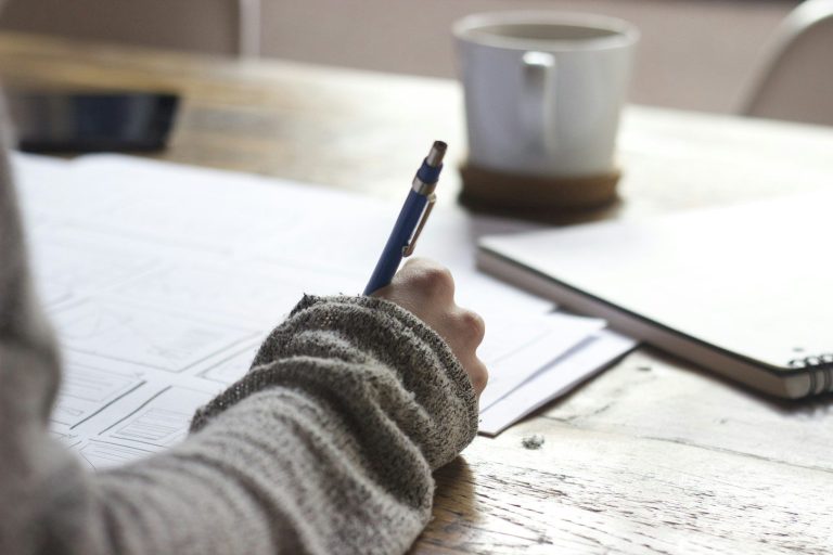 A person holding a pen, writing on paper, with a notebook to work out ways to save money and a cup of coffee nearby.